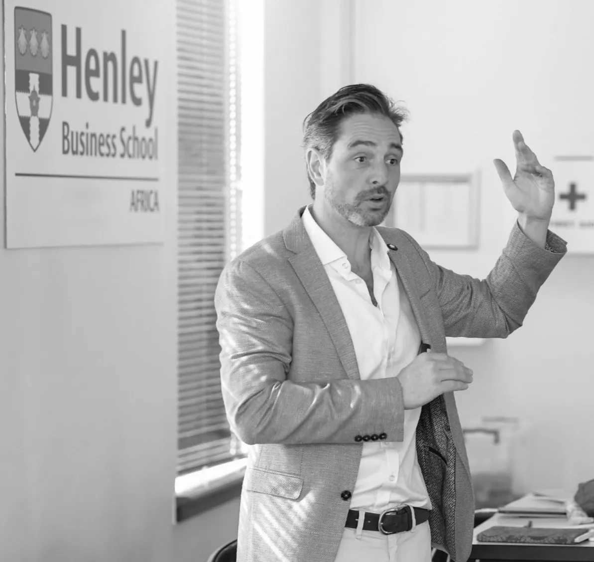 David Joshua mid-sentence, one hand raised, in a light blue jacket and open-necked white shirt, beside the Henley Business School Africa sign