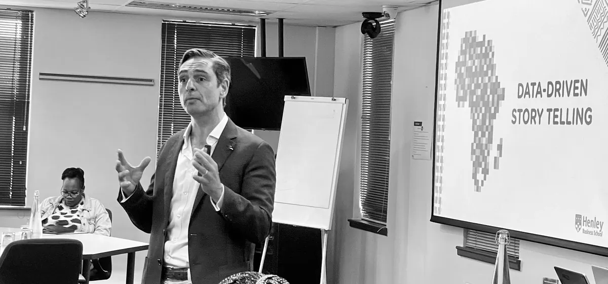 David Joshua teaching, mid-gesture, in a seminar room; beside him a screen reads Data-Driven Story Telling over a map of Africa, with the Henley Business School mark