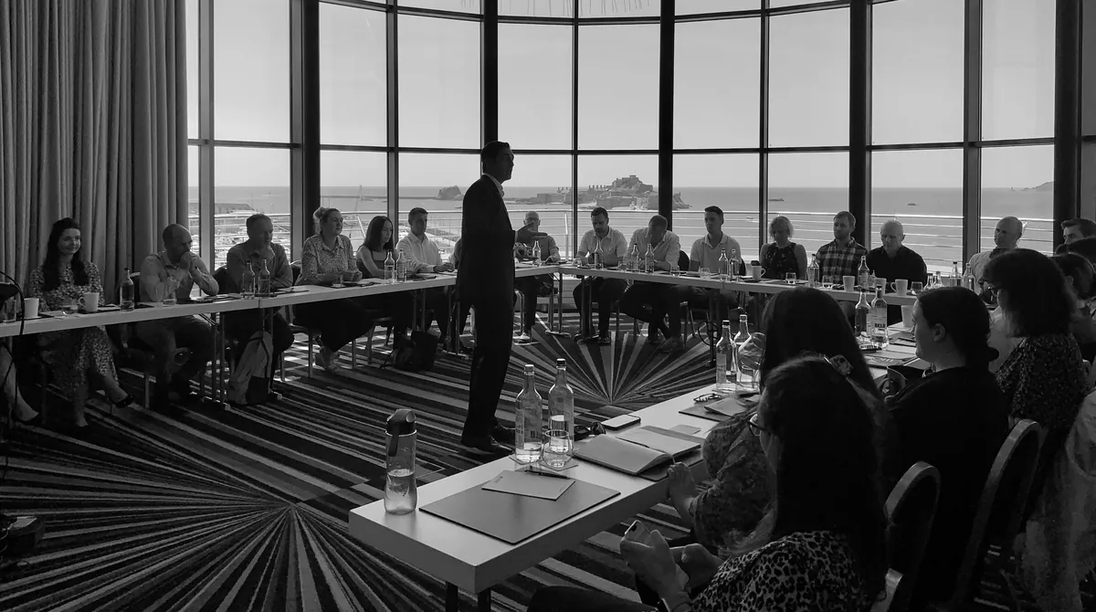 David Joshua standing at the head of a U-shaped conference table, addressing about twenty delegates, with the sea and an island fort through the floor-to-ceiling windows behind him