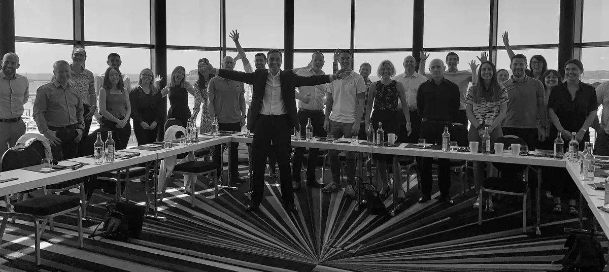 A cohort of about thirty people standing around a U-shaped table with their arms raised, David Joshua at the centre, in a round room with floor-to-ceiling windows and the sea behind